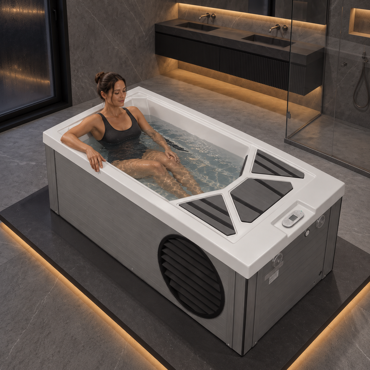 A woman relaxes in a white and gray heated outdoor bathtub filled with water, set on a dark platform in a modern bathroom with warm under-cabinet lighting.