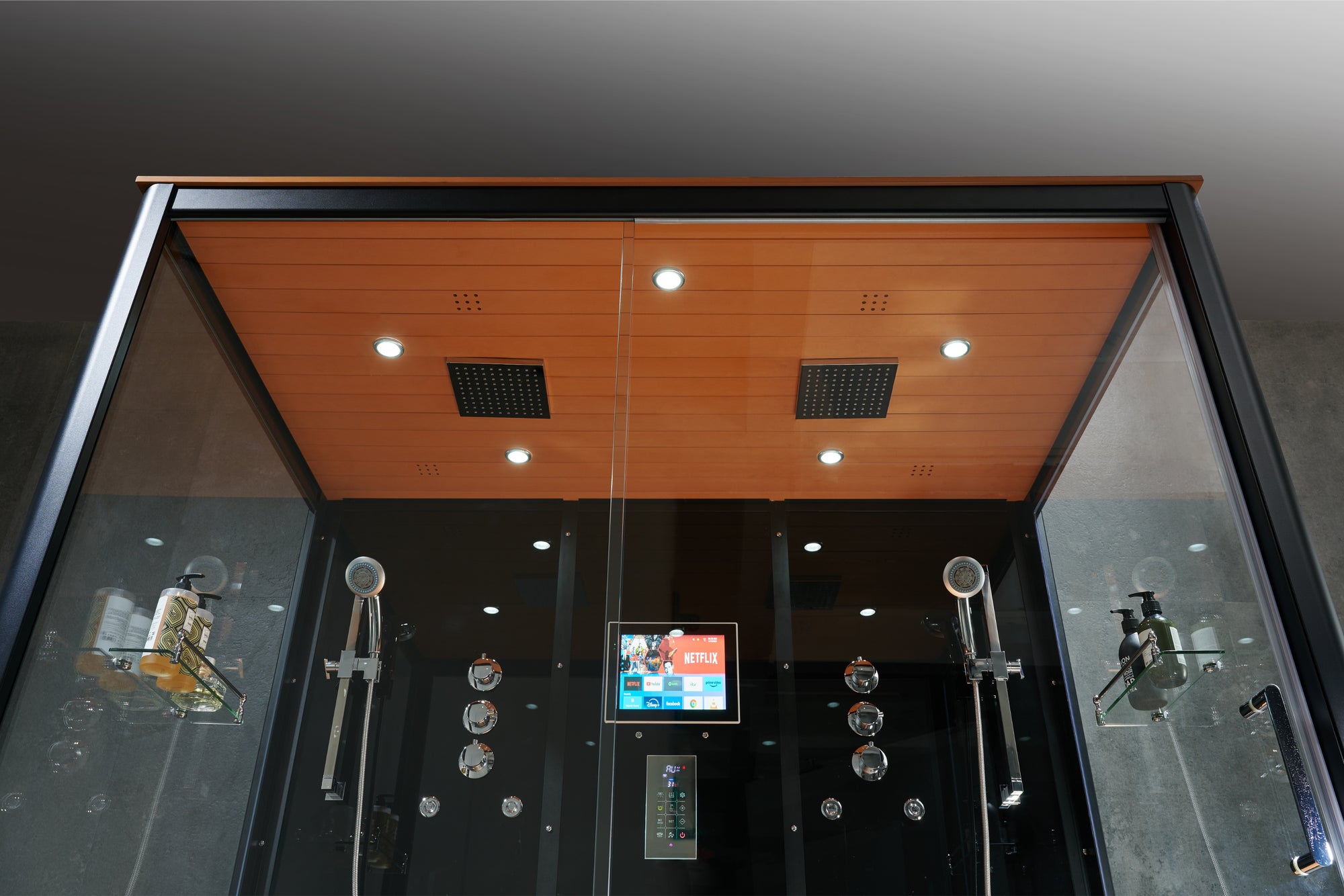 A modern large-format rain shower cabin with a black frame and glass enclosure, featuring a wood-look ceiling, built-in LED lighting, dual handheld showerheads, and a central digital control screen.
