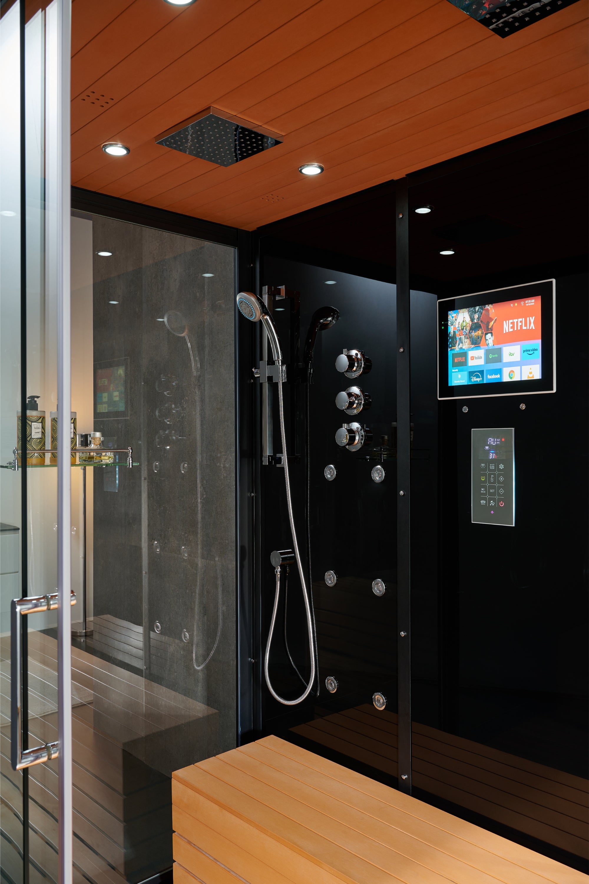 A modern indoor hot tub or steam shower enclosure with a black control wall, wood ceiling and bench, glass enclosure door, and an on-screen media display.
