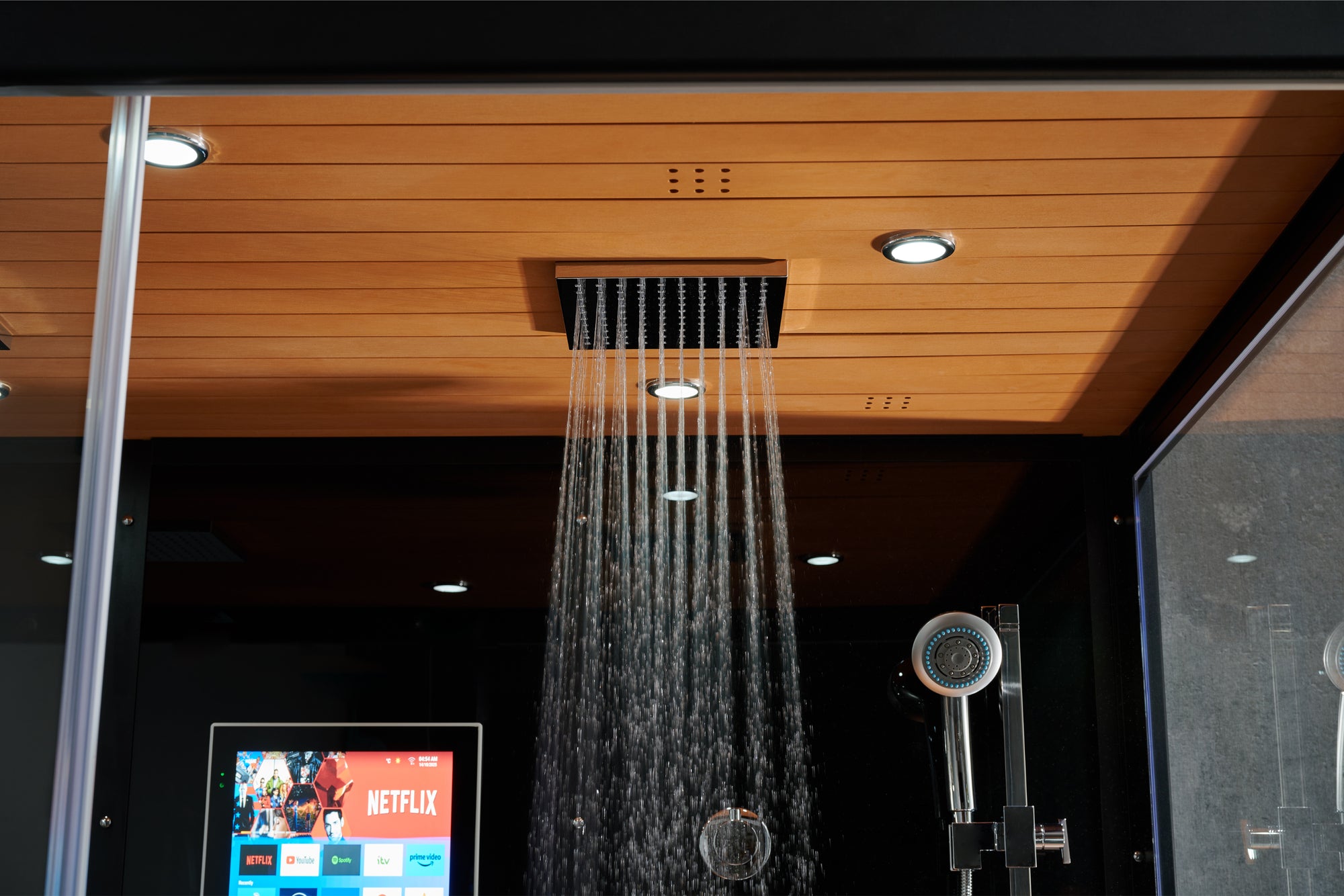 A modern rectangular rain shower head mounted on a wooden ceiling, with water spraying downward. The shower is installed in a bathroom or shower enclosure with dark walls, recessed lighting, and a handheld shower fixture visible to the right.