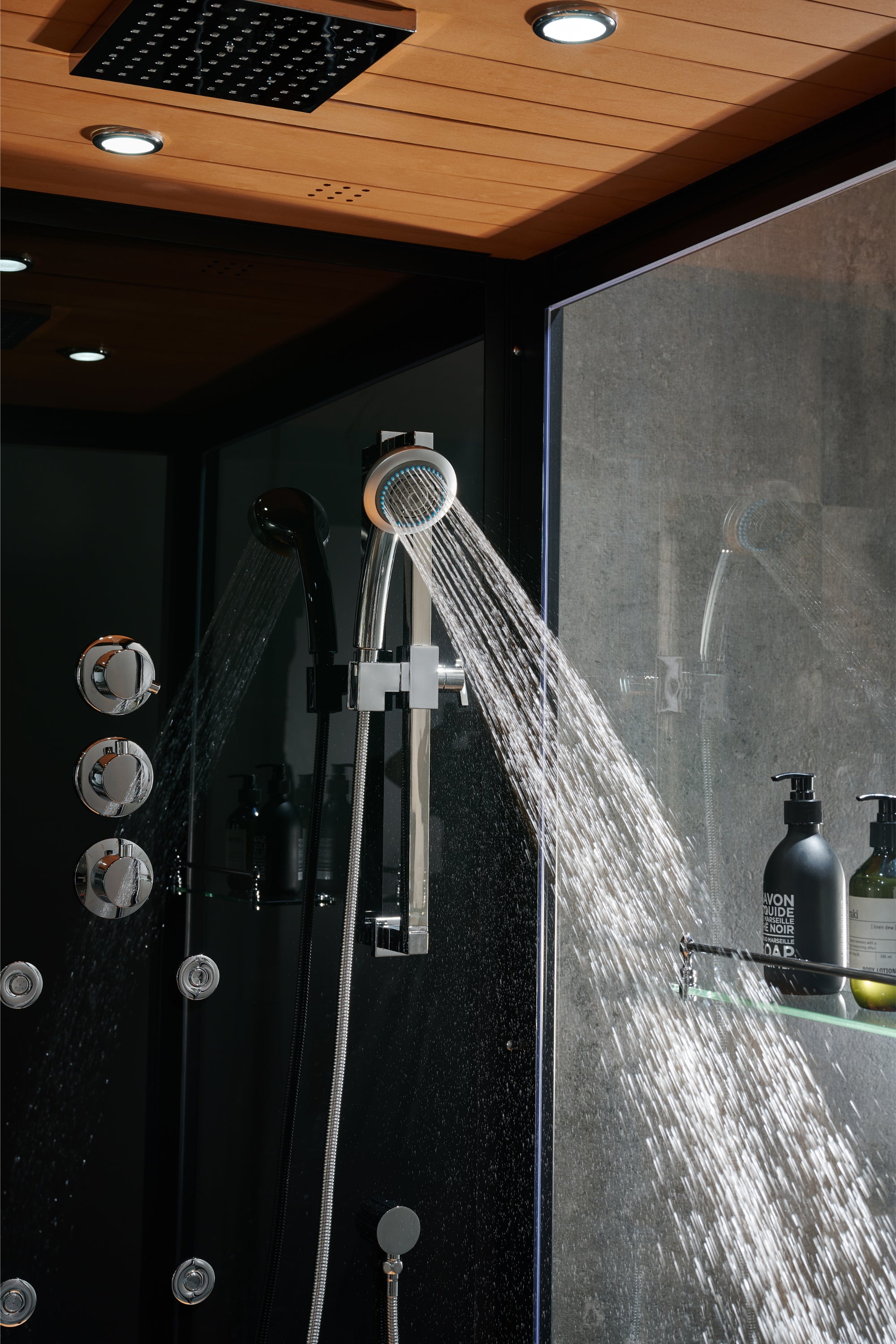 A modern shower system with a handheld showerhead spraying water and a rain shower head overhead, set in a dark bathroom with wood ceiling panels and a glass enclosure.