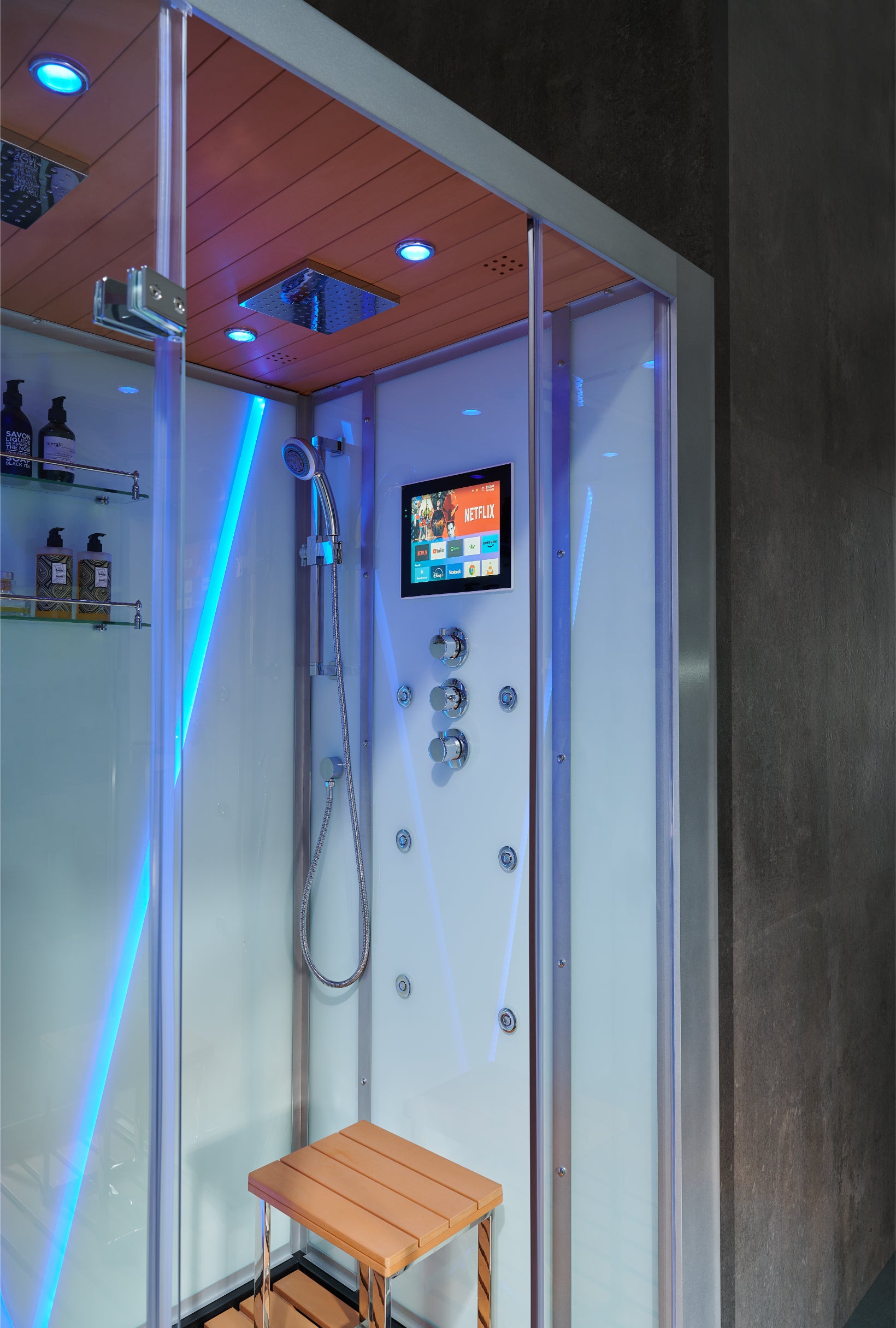 A modern glass shower cabin with a wooden ceiling, blue LED lighting, a built-in control screen, handheld shower, and a small wooden bench inside.