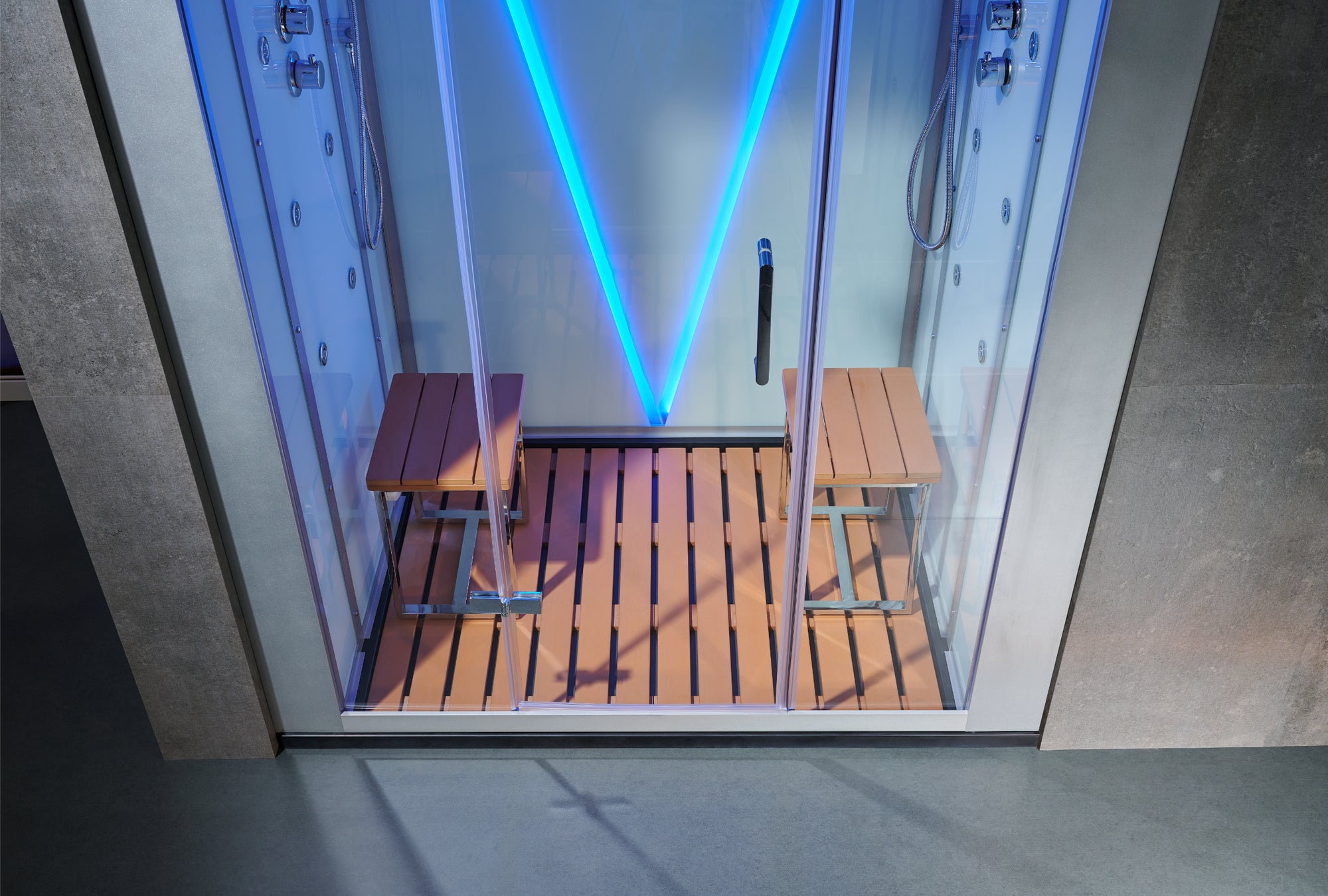 A glass steam room or sauna with a wooden slatted floor, two wooden benches, and bright blue LED lighting inside, shown against a gray concrete wall.