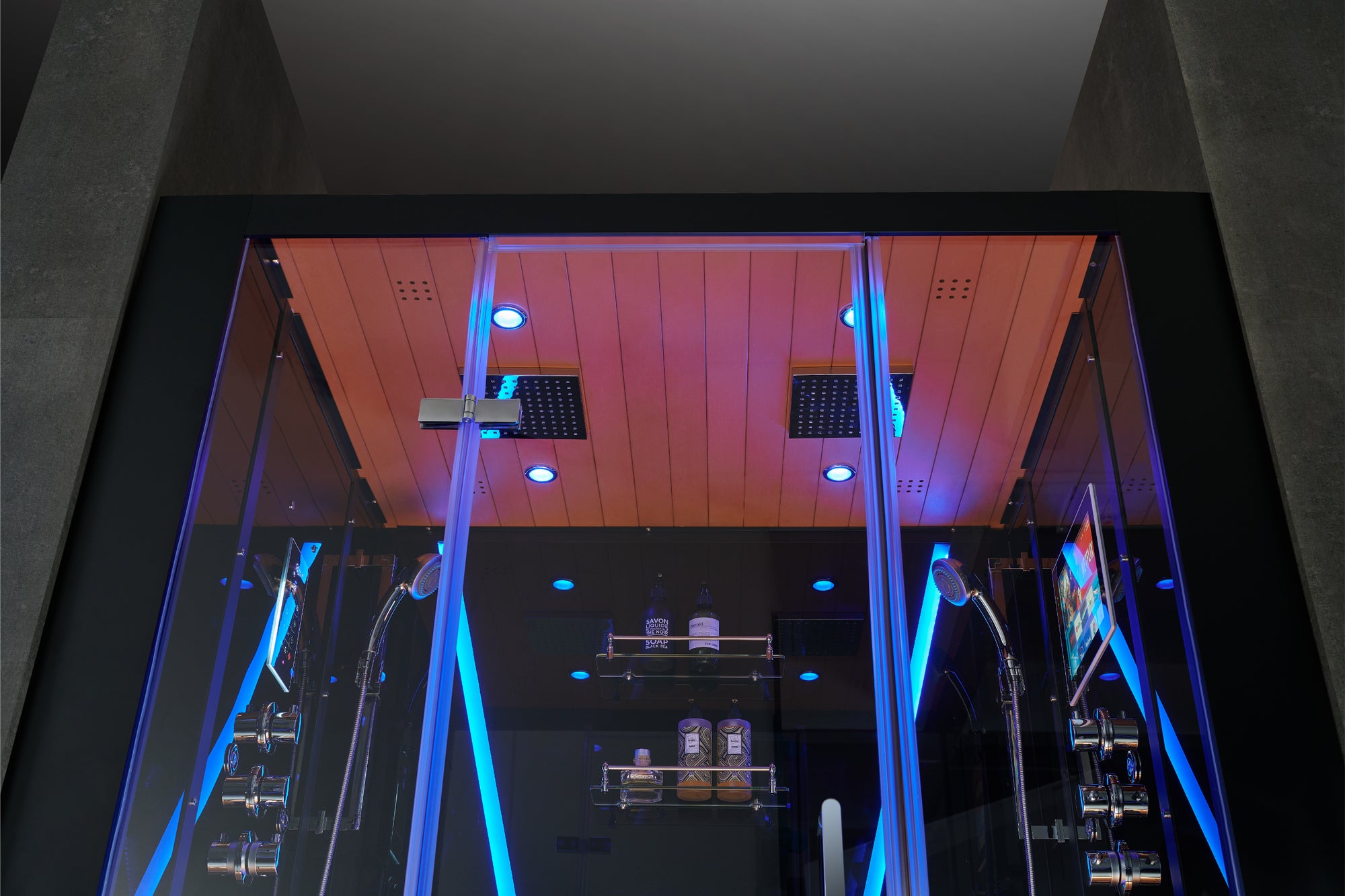 A modern glass rain shower cabin with blue LED lighting accents, featuring a wooden ceiling panel and built-in shower heads visible through the glass doors.