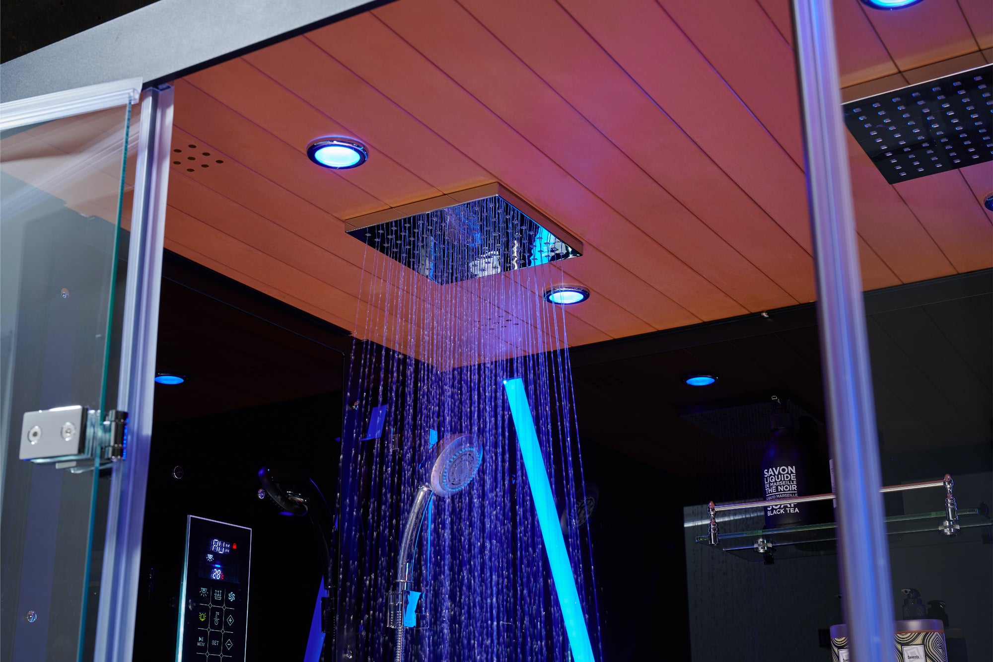 A modern rain shower system installed in a glass shower enclosure, featuring a square ceiling-mounted shower head with water spraying down, illuminated by blue LED lighting. The shower room has a wood-look ceiling panel and recessed lights.