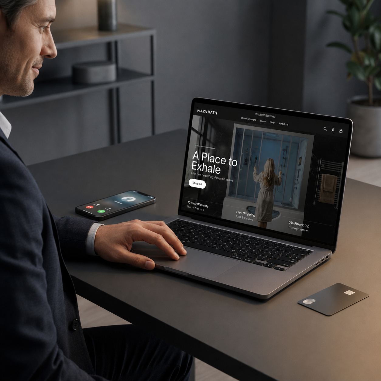 A man in a dark suit sits at a desk using a laptop that displays a website for 'Maya Bath', with a smartphone and a credit card on the table in a modern office setting.